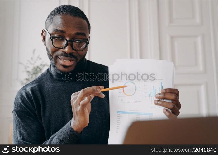 Pleasant immersed in work African male home office worker holds business meeting online with coworkers, holding in hand sales charts and pointing with pencil important factors during video call. Pleasant immersed in work African male home office worker holds business meeting online