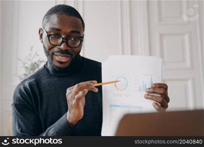 Pleasant immersed in work African male home office worker holds business meeting online with coworkers, holding in hand sales charts and pointing with pencil important factors during video call. Pleasant immersed in work African male home office worker holds business meeting online