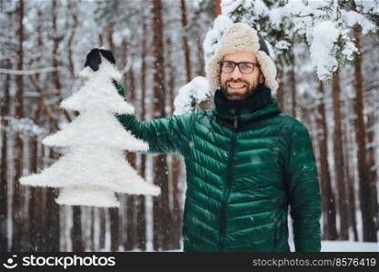 Pleasant looking bearded male dressed in warm clothes, holds artificial fir tree, stands against beautiful trees covered with snow, has cheerful positive expression, breathes fresh winter air