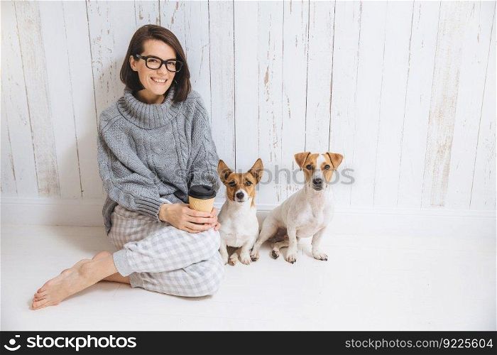 Pleasant looking brunette female dressed casually, drinks hot beverage from paper cup, sits near two dogs, enjoys domestic atmosphere, looks directly into camera. People, leisure, pets concept