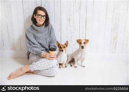 Pleasant looking brunette female dressed casually, drinks hot beverage from paper cup, sits near two dogs, enjoys domestic atmosphere, looks directly into camera. People, leisure, pets concept