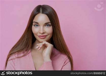 Pleasant looking brunette female fashion model keeps hand under chin, has full lips and applealing appearance, wears rosy jumper in one tone with background. Women, cosmetic and skin care concept