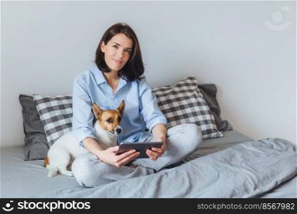 Pleasant looking brunette female in pyjamas uses modern electronic gadget in bedroom on bed, sits with her jack russell terrier dog, enjoys calm domestic atmosphere, has nice weekends