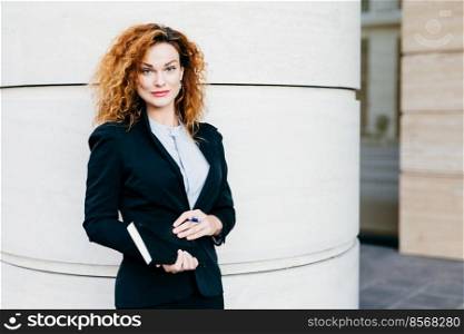 Pleasant-looking curly woman dressed formally, holding notebook in hands, going to have meeting wit her business partners. Gorgeous business lady writing in diary book. Professionalism concept