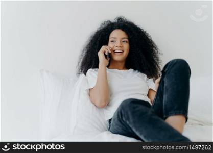 Pleasant looking curly woman has pleasant conversation, talks with friend via smartphone while relaxes in bed, has joyful expression, looks aside, wears casual clothing. People, bedding, technology