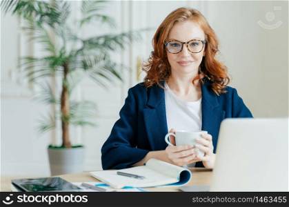 Pleasant looking elegant female freelancer books items on web store, reads news in internet, writes notes in notepad, works on laptop computer, dressed in elegant outfit. Business and work concept