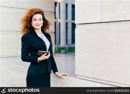 Pleasant-looking female dressed formally, holding digital tablet computer which is necessary for her work. Beatiful woman with curly bushy hair, having attractive appearance, going for her work