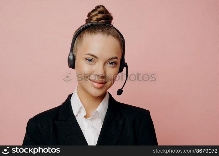 Pleasant looking female office worker in dark suit with black wireless headset communicating with coworkers online trough web conference, looking with smile at camera isolated over pink background. Pretty business lady in headset being happy and satisfied after online meeting
