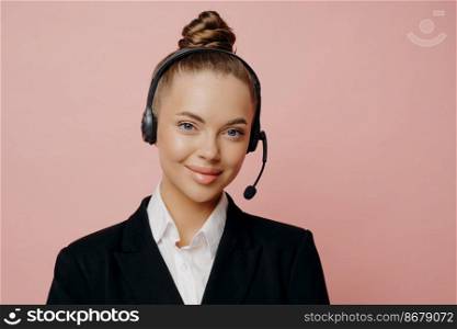 Pleasant looking female office worker in dark suit with black wireless headset communicating with coworkers online trough web conference, looking with smile at camera isolated over pink background. Pretty business lady in headset being happy and satisfied after online meeting