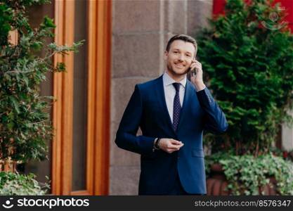 Pleasant looking glad businessman has conversation on smart phone, calls partner as discuss financial problems of company, happy to hear colleague, uses modern technologies, dressed in formal suit