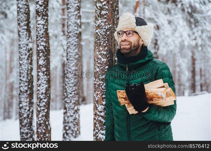 Pleasant looking happy male holds firewood, looks thoughtfully aside, stands near winter trees, dreams about something pleasant, enjoys beautiful winter landscapes. Nature concept.