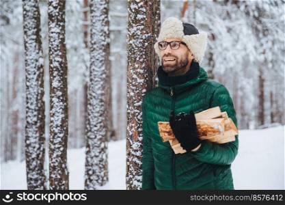 Pleasant looking happy male holds firewood, looks thoughtfully aside, stands near winter trees, dreams about something pleasant, enjoys beautiful winter landscapes. Nature concept.