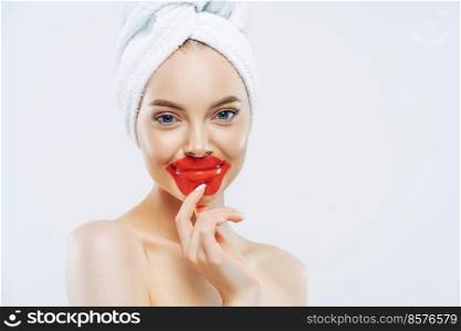 Pleasant looking healthy woman keeps patches on lips, wears towel on head, has clean pure skin after hygienic procedure, stands shirtless against white background, empty space for your advertisement