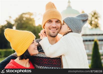 Pleasant looking little girl being thankful to her father for buying new bicycle, kisses him in cheek. Young family walk together outdoor, spend spare time in park, wear knitted fashionable hats