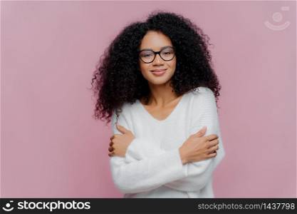 Pleasant looking millennial Afro American woman keeps hands crossed over chest, has curly bushy hair, wears white comfortable sweater and spectacles, isolated over purple background, feels pleased