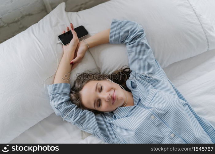Pleasant looking relaxed young woman has satisfied expression, dreams about something, wears casual clothes, lies on white pillow, connected to smart phone, listens nice music, has rest in bed