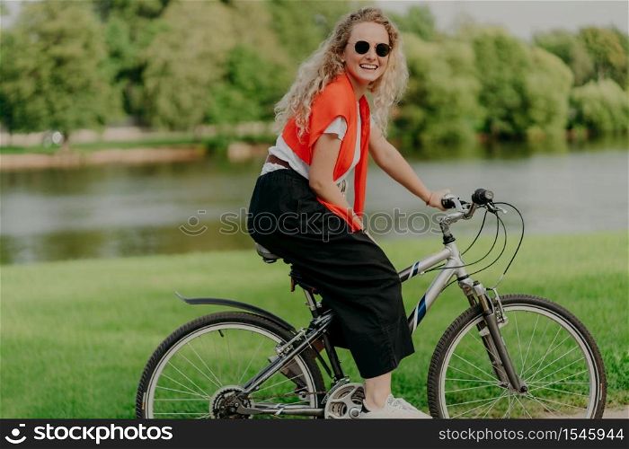 Pleasant looking satisfied curly haired woman spends free time in open air, rides bicycle, spends leisure alone, enjoys freshness of nature, looks at camera with pleasant smile. Summer time.