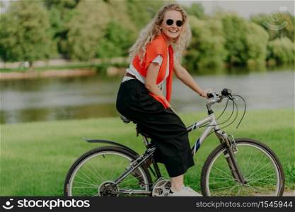 Pleasant looking satisfied curly haired woman spends free time in open air, rides bicycle, spends leisure alone, enjoys freshness of nature, looks at camera with pleasant smile. Summer time.