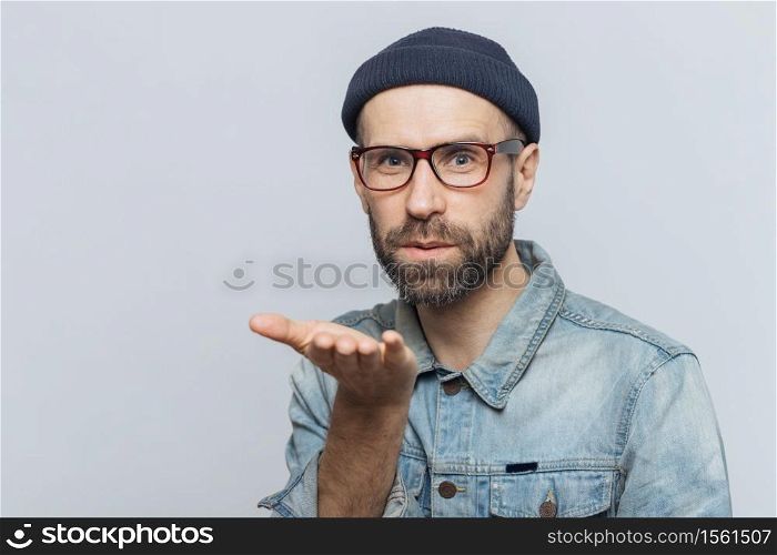 Pleasant looking young bearded male with mustache, wears fashionable glasses, black hat and denim shirt, blows air kiss at camera, expresses his love to girlfriend, isolated over grey background