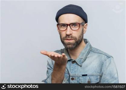 Pleasant looking young bearded male with mustache, wears fashionable glasses, black hat and denim shirt, blows air kiss at camera, expresses his love to girlfriend, isolated over grey background