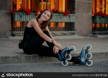 Pleasant looking young European woman wears rollerskates takes break after riding poses outdoor dressed in black activewear has happy smile on face. People recreation hobby and lifestyle concept