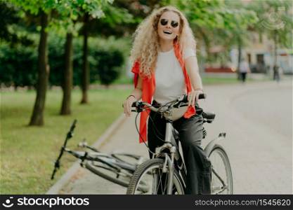 Pleasant looking young woman in sunglasses, rides sport bicycle, stops for break, enjoys beauty of nature, calm atmosphere, laughs positively, keeps both hands in handlebars, discovers new place