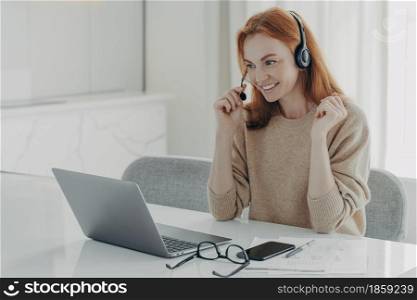 Pleasant smiling redhead woman in headset communicating with colleagues during video call on laptop computer, female freelancer participating in online meeting while working remotely from home. Pleasant smiling redhead woman in headset communicating with colleagues during video call on laptop