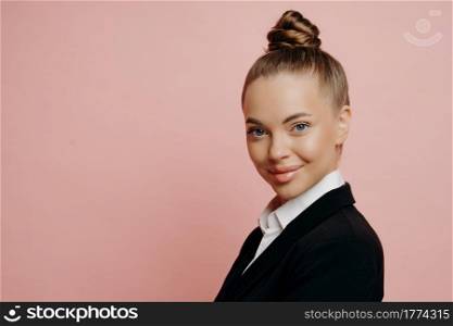 Pleasant woman in classic black suit looking happily and gladfully at camera, being professional in her field of work, isolated over pink background. Studio shot of successful business woman. Female business professional posing against pink wall in studio