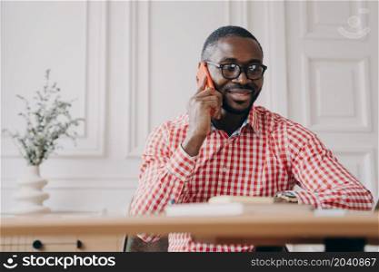 Pleasant young african american businessman talking on mobile phone and consulting client while working in office, smiling dark-skinned male office worker in shirt making call on smartphone. Pleasant young afro american businessman talking on mobile phone with client, working in office