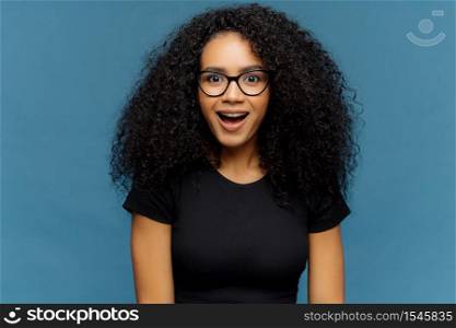 Pleasantly surprised dark skinned woman keeps jaw dropped, gazes with interest, has curly hair, wears black t shirt, models against blue wall. Afro American lady gazes with shock, has bated breath