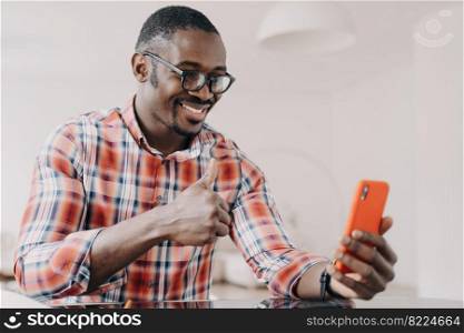 Pleased african american man showing thumb up hand gesture, holding smartphone, chatting by video call. Smiling black guy in glasses looking at phone screen, shows like sign, sitting at desk.. Pleased african american man shows thumb up hand gesture, holding phone, chatting by video call