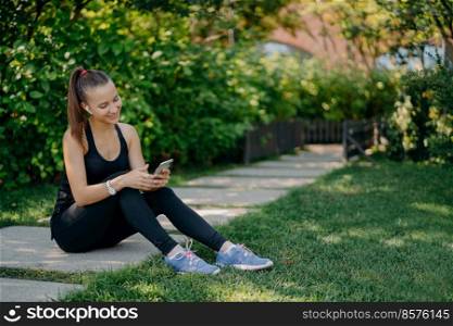 Pleased athletic woman in sportsclothes smiles cheerfully surfs mobile phone while listens music via wireless earphones takes break after sunny morning workout uses app for activity tracking