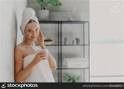Pleased attractive woman has natural beauty, glowing smooth healthy skin, touches face, enjoys spa procedures, wears white bath towel on head and around body, drinks tea, poses in cozy bathroom