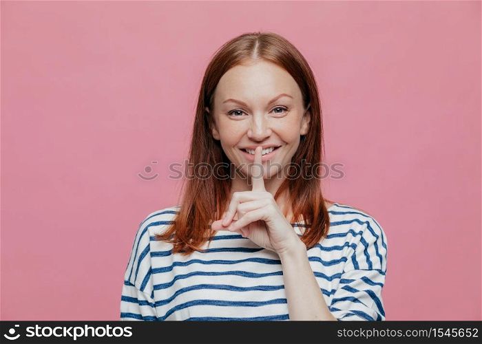 Pleased attractive woman with brown hair, gentle smile, keeps index finger on lips, shows silence gesture, dressed in striped clothes, models over pink background. People and conspiracy concept