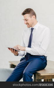 Pleased attractive young businessman checks email on portable pc, watches vidio on internet webpage, uses free internet connection, wears elegant formal clothes, isolated over white background