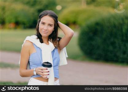 Pleased beautiful European woman with fit figure, keeps hand on head, drinks takeaway coffee, stands against green blurred background with copy space for your advertisement. Coffee time concept