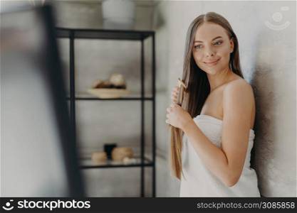 Pleased beautiful female model combs her long hair with hairbrush, stands in bathroom wrapped in soft towel, has well groomed complexion and healthy skin. Hair care and beauty routine concept