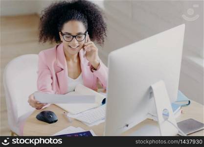Pleased beautiful gorgeous curly haired businesswoman looks through documents, has telephone talk, sits at worplace, uses computer for making financial report, wears spectacles for vision correction