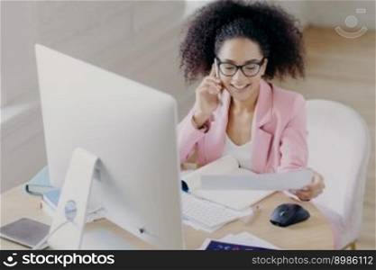 Pleased beautiful gorgeous curly haired businesswoman looks through documents, has telephone talk, sits at worplace, uses computer for making financial report, wears spectacles for vision correction