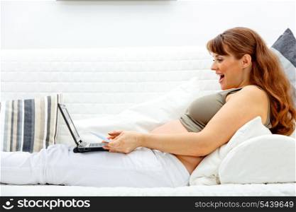 Pleased beautiful pregnant woman sitting on sofa at home with laptop and credit card&#xA;