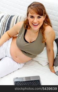 Pleased beautiful pregnant woman sitting on sofa at home with laptop&#xA;