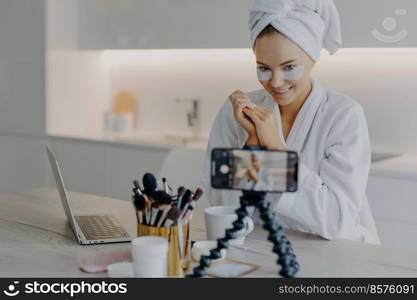 Pleased beauty blogger sits in front of cellphone camera shares experience about skin care wears hydrogel patches under eyes poses at table with cosmetic tools wears bathrobe wrapped towel on head