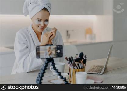 Pleased beauty blogger sits in front of cellphone camera shares experience about skin care wears hydrogel patches under eyes poses at table with cosmetic tools wears bathrobe wrapped towel on head