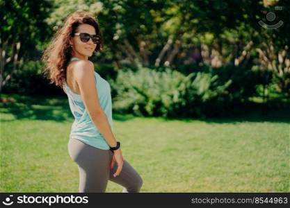 Pleased brunette European woman in active wear poses sideways wears sunglases smartwatch has fitness outdoors rests after jogging smiles pleasantly. Health body care sport and exercising concept