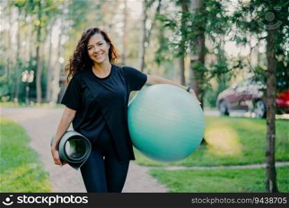 Pleased brunette holds rolled karemat and fitness ball, dressed in black t-shirt. Prepares for aerobic exercises, smiling against nature backdrop. Sport concept.