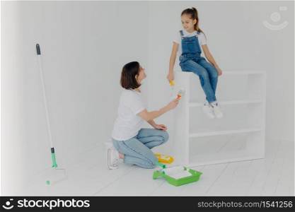 Pleased brunette mother looks at child, paint furniture in modern apartment, poses in spacious empty room with white walls, busy doing home renovation. Family, repairing and painting concept