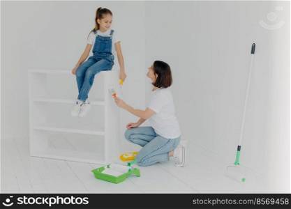 Pleased brunette mother looks at child, paint furniture in modern apartment, poses in spacious empty room with white walls, busy doing home renovation. Family, repairing and painting concept