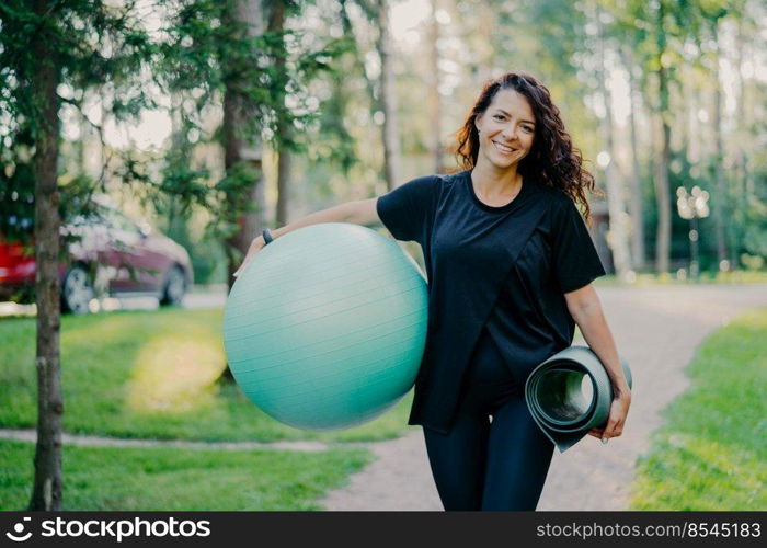 Pleased brunette woman dressed in black t shirt, holds rolled up karemat and fitness ball, prepares for aerobic exercises, smiles pleasantly, poses against nature background. People and sport concept