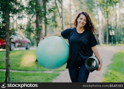 Pleased brunette woman dressed in black t shirt, holds rolled up karemat and fitness ball, prepares for aerobic exercises, smiles pleasantly, poses against nature background. People and sport concept
