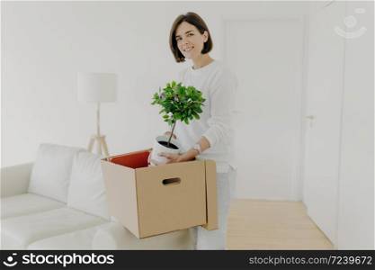 Pleased brunette young woman unpack belongings in carton box, holds green plant in pot, relocates in new apartment, dressed in casual clothes, stands in spacious room, being busy with house work